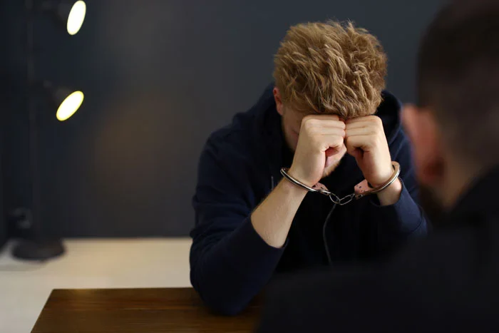 A young man in handcuffs sits at a table, his head in his hands, expressing distress during a serious conversation